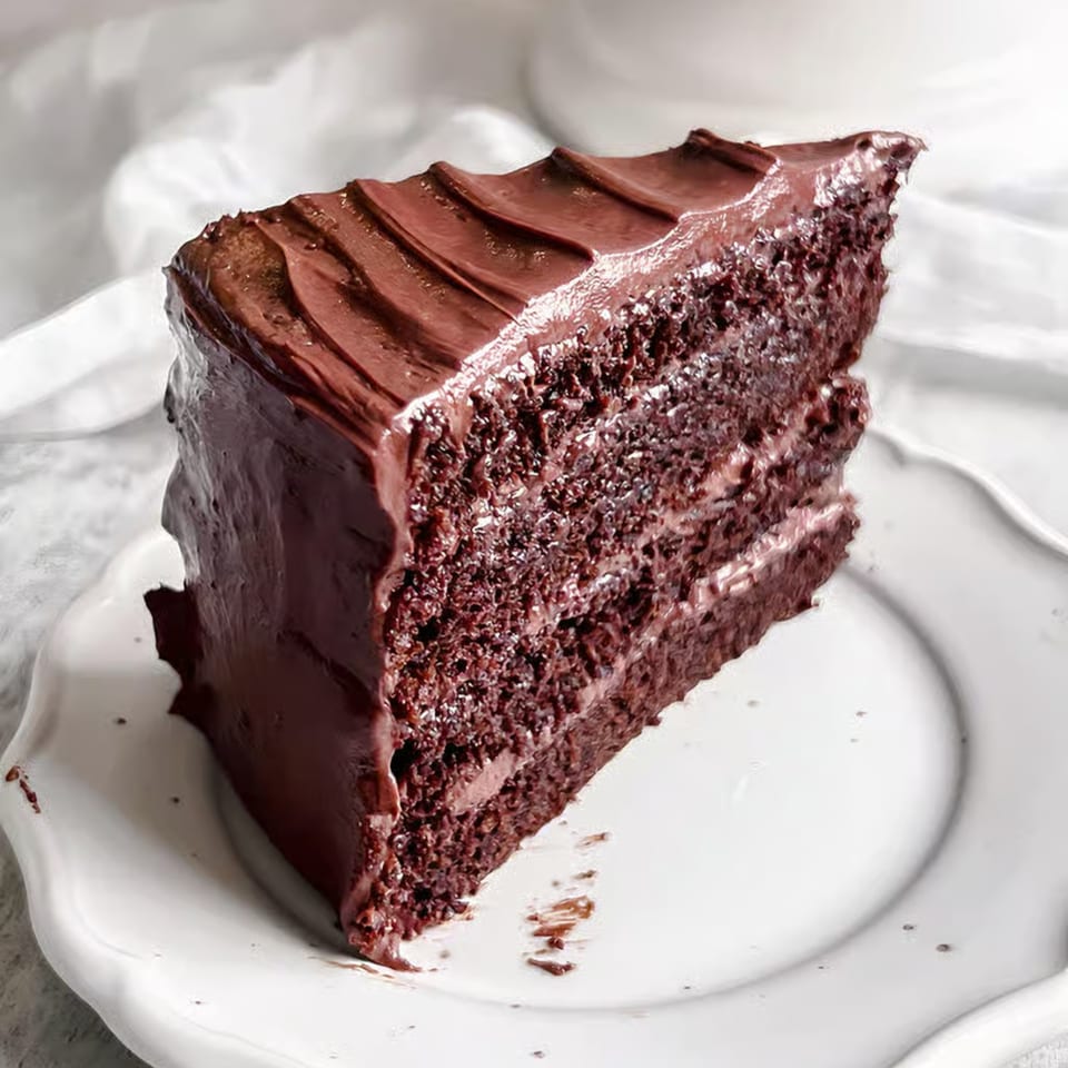 A rich slice of chocolate cake with thick chocolate frosting on a white plate.
