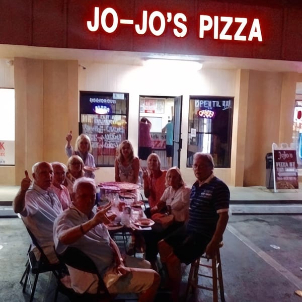 A group of people enjoying pizza outside Jo-Jo's Pizza restaurant at night.