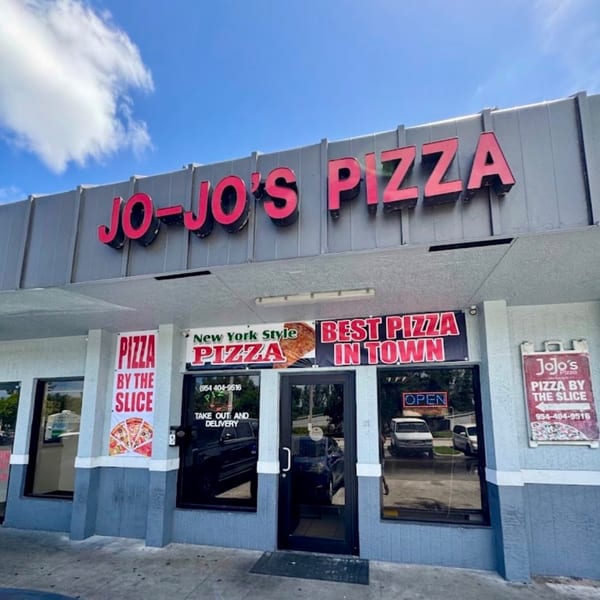 The exterior of Jo-Jo's Pizza, featuring a red sign and window advertisements for New York-style pizza.